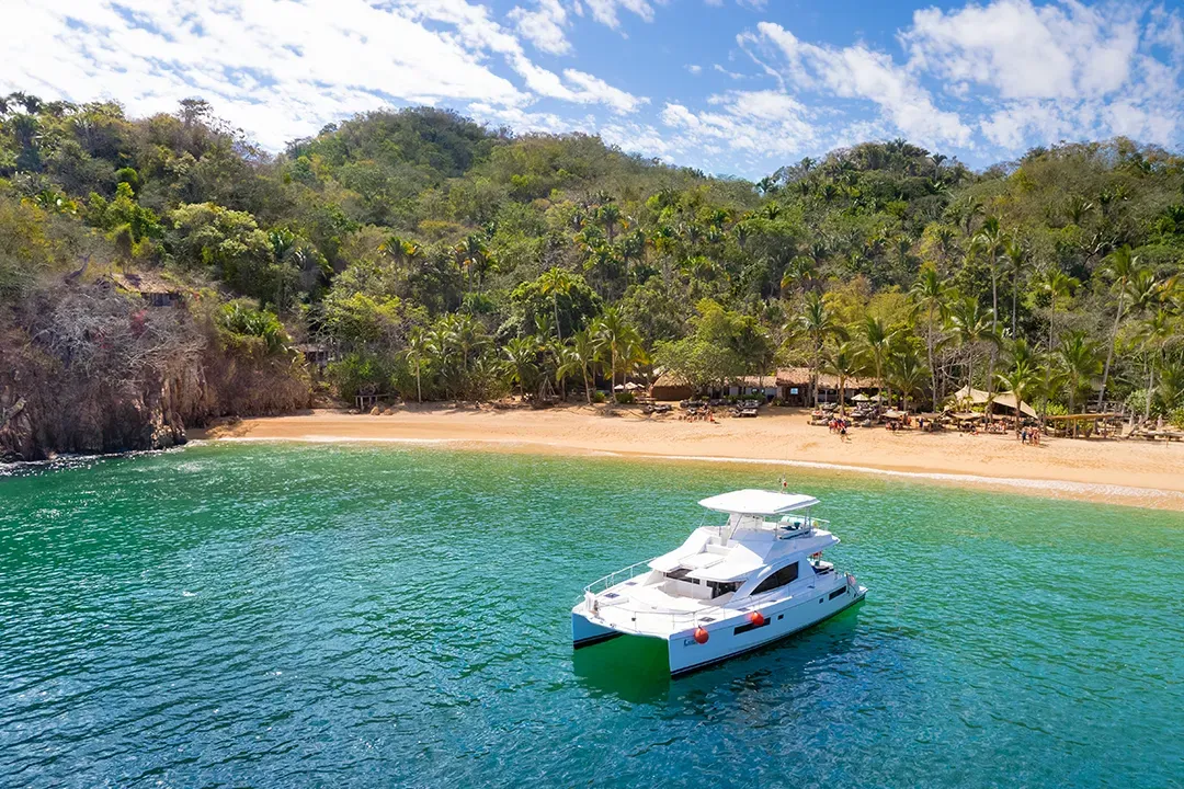 A modern white catamaran sailing near Los Arcos Marine Park with the green hills of Puerto Vallarta in the background.